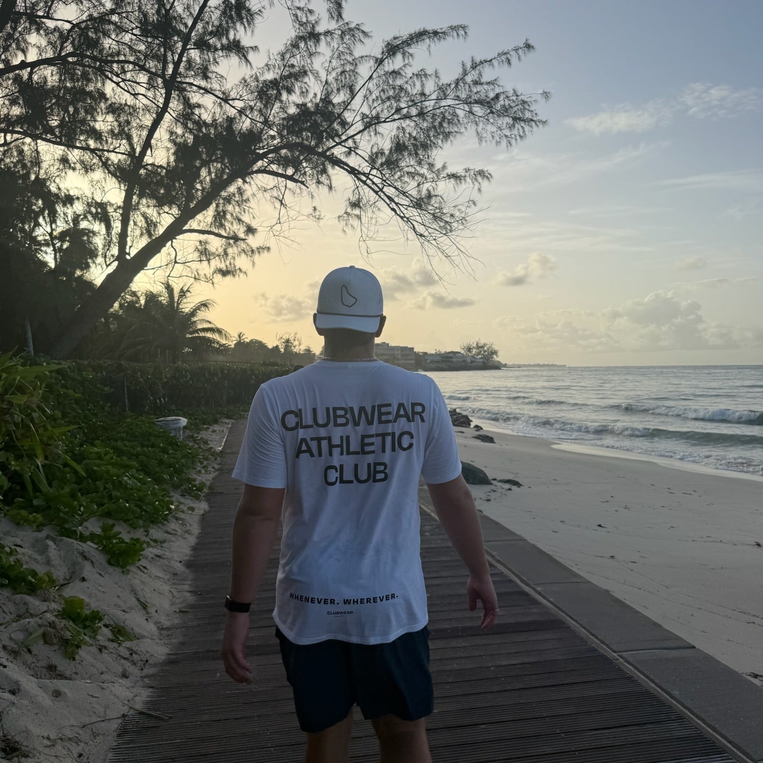 Person walking on a wooden path by the beach wearing a 'Clubwear Athletic Club' t-shirt.
