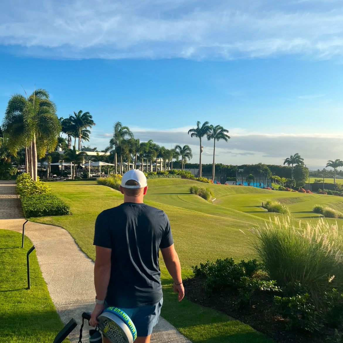 Man walking with a golf bag on a golf course with palm trees and a clear sky.