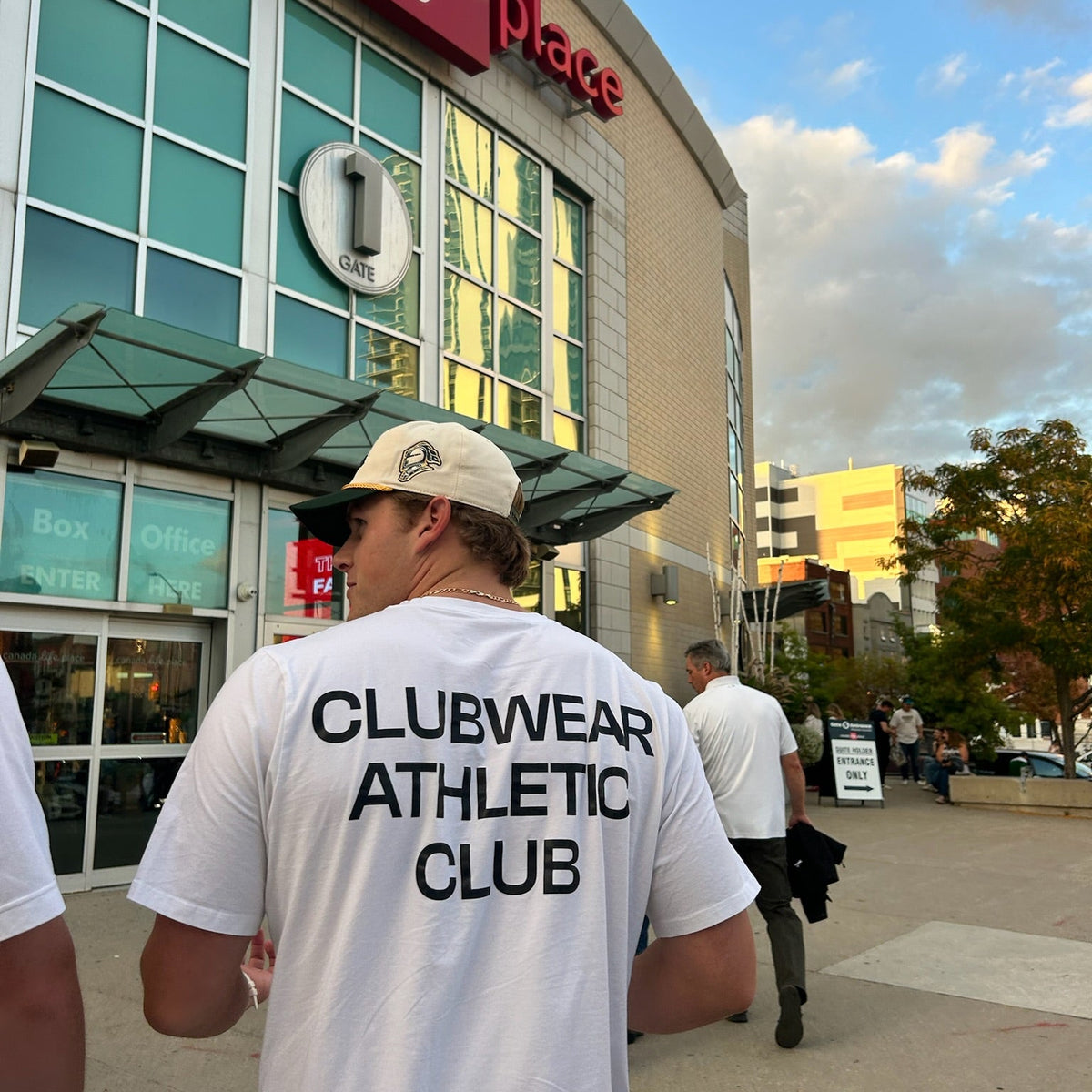 Man wearing a 'Clubwear Athletic Club' t-shirt walking past a store with a visible brand logo.